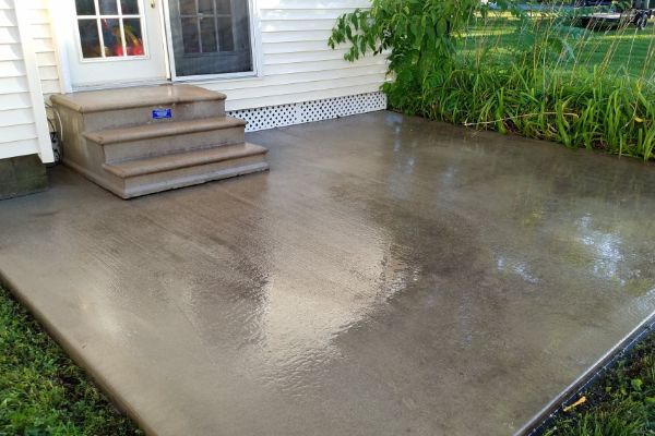 After: A freshly cleaned concrete patio is shown outside a white house, next to a set of three steps leading to a door. The surface appears wet and glossy, with green plants along the edge.