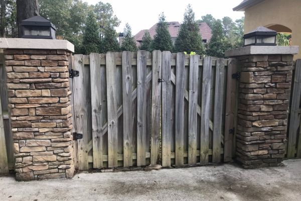 Before: A wooden fence gate with vertical planks and two stone pillars on each side, located on a concrete surface, with trees and a house visible in the background.