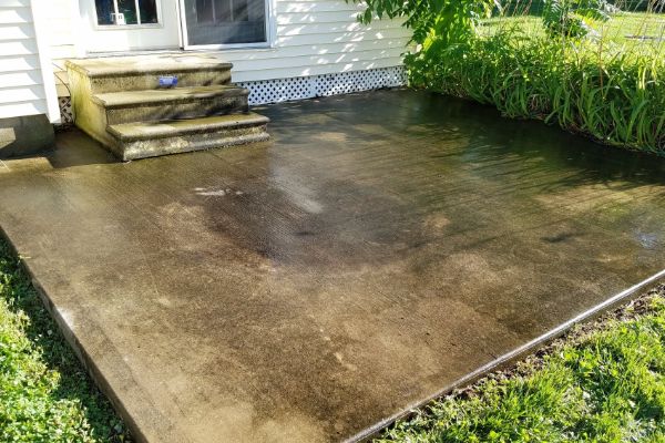 Before: A wet, but dirty concrete patio outside a house, with steps leading to a door and green plants bordering one side. Sunlight creates reflections on the clean surface.