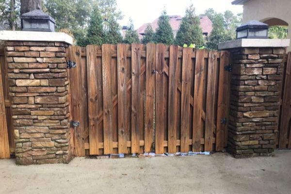 After: A wooden fence gate with vertical planks stands between two stone pillars on a concrete driveway, with trees and a house visible in the background.