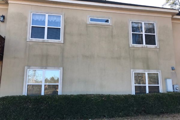 Before: A beige stucco house exterior with two pairs of double windows on the lower level, two pairs of double windows and a small horizontal window above, showing dark vertical streaks of dirt or mildew on the wall. Shrubs line the base.