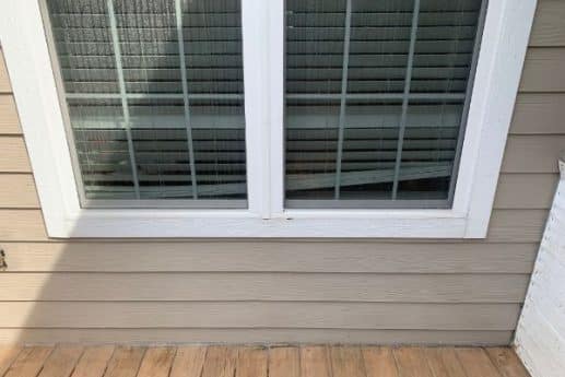 After: A double-paned window with white trim is set in beige horizontal siding that has been cleaned by a soft washing service. A wooden deck runs along the bottom edge.