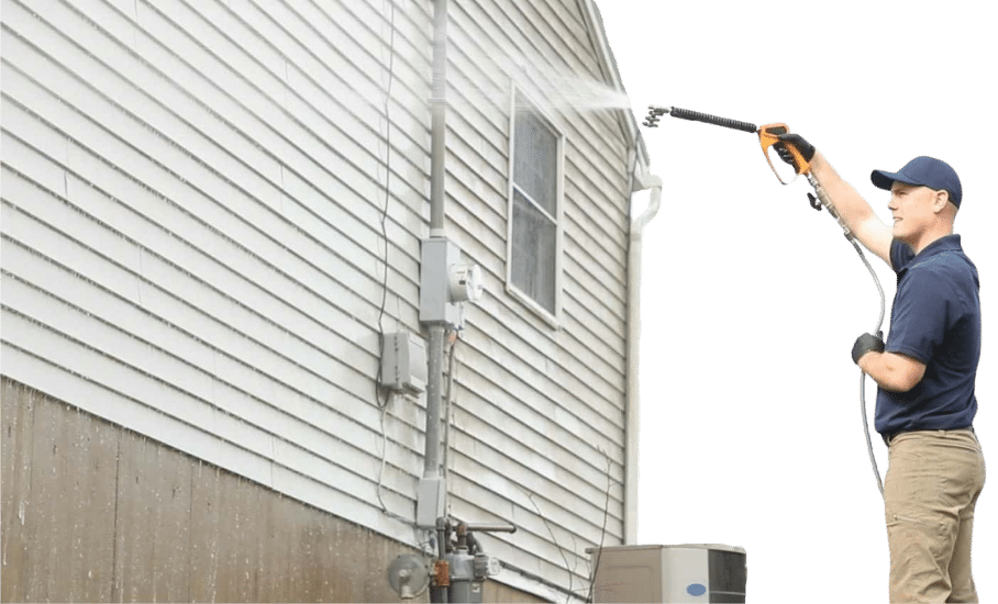 A man wearing gloves and a cap uses a power washer for house washing, cleaning the exterior siding and spraying water upward toward a window.