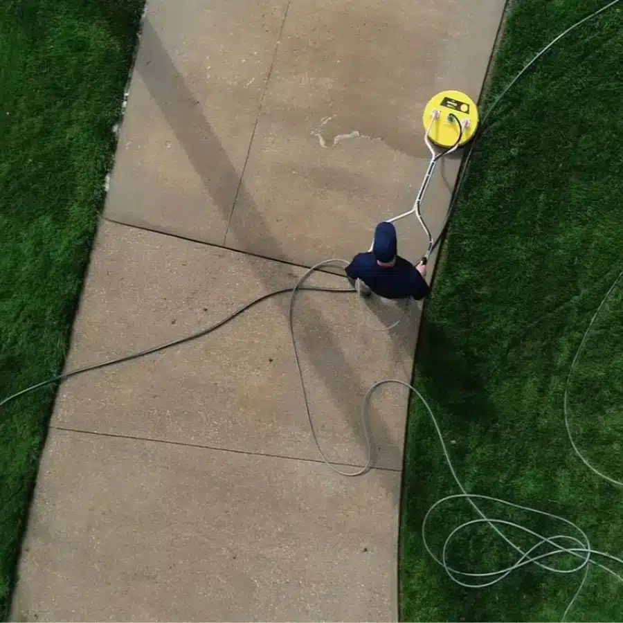 A person operates a pressure washer on a concrete sidewalk, with green grass nearby and hoses laid out on the ground.